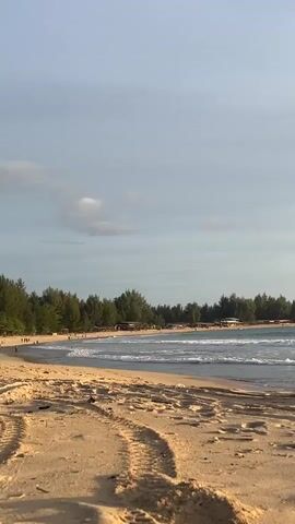 This is a wide shot of a sandy beach in Meunasahmesjidlampuuk, Indonesia, at what appears to be late afternoon or early evening due to the warm lighting and long shadows cast on the sand. The sky is a pale blue with a few wispy clouds. Gentle waves are rolling onto the shore, creating a serene atmosphere.

In the distance, a line of trees, likely pine or similar, borders the beach. Behind the trees, several low-lying buildings with dark roofs are visible, suggesting a small resort or residential area. A few people are scattered along the shoreline and in the shallow water, some appearing to be walking or standing. The activity is calm and leisurely, characteristic of a relaxed beach setting.

The foreground is dominated by the textured sand, with distinct tracks suggesting recent human or possibly animal activity. The overall mood is tranquil and peaceful, with the sounds of the ocean likely being prominent. There is no visible text in the image.