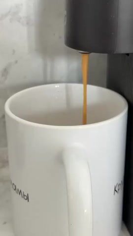 A close-up, slightly angled shot shows a coffee machine dispensing a stream of dark liquid into a white ceramic mug. The liquid, likely coffee or espresso, flows from a narrow spout on the machine into the mug, creating ripples on the surface. The mug has a handle and appears to have some text in black, partially visible on its side. The background consists of a marbled surface and a wall with a light, textured pattern. The lighting is bright and even, highlighting the liquid and the objects. The video is very short, capturing only a few seconds of the coffee-making process.