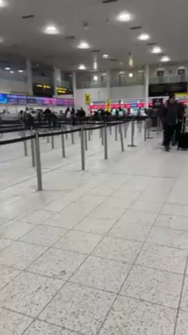 The video shows the inside of an airport.  The floor is made of square tiles, and there are metal stanchions set up to guide people.  Several people are walking through the area, some pulling luggage behind them.  In the background, there are check-in counters with lights above them. The room is well-lit.