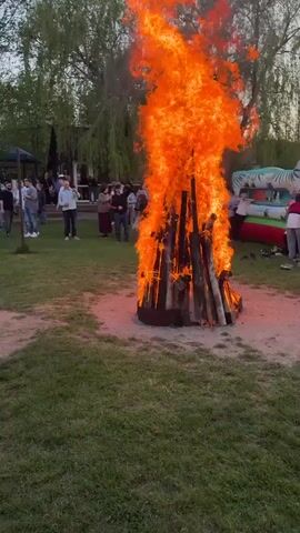 The video captures a gathering of people in an outdoor setting with trees in the background. Initially, the focus is on a group of people standing on a grass field. Subsequently, the scene shifts to a large bonfire with tall flames in the center of the field. People are standing around the bonfire, and an inflatable bounce house is visible nearby. A man with a beard and a hat is standing and seemingly directing the group. A few strollers and children's trikes are scattered around the field. The day appears to be late afternoon or early evening with ambient light.