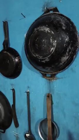 The video shows a collection of kitchen utensils hanging on a blue wall. The utensils include various pots, pans, ladles, and spatulas. Some of the items appear to be made of metal, while others have wooden or plastic handles. The wall is painted a bright blue and appears to be somewhat worn. There are also a few nails or hooks visible on the wall from which the utensils are suspended. The lighting in the video is dim, suggesting it may be an indoor setting with limited natural light. In the foreground, there is a glimpse of a wooden structure on the left side of the frame, possibly part of a shelf or cabinet.