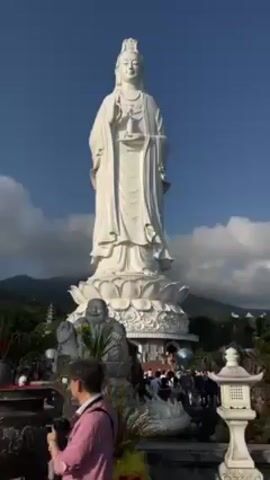 The video shows a large white statue of a woman in front of a blue sky with white clouds. There is a crowd of people near the base of the statue and some smaller statues as well. A man in a pink shirt is taking a picture of the statue. The video then pans up to show a tree with red ribbons tied to the branches. There are a few people standing near the tree, including a woman with blonde hair and a bald man. The video then shows a view from a high vantage point, looking out over a body of water towards a city skyline. The sun is shining brightly and reflecting off the water. There are trees in the foreground.