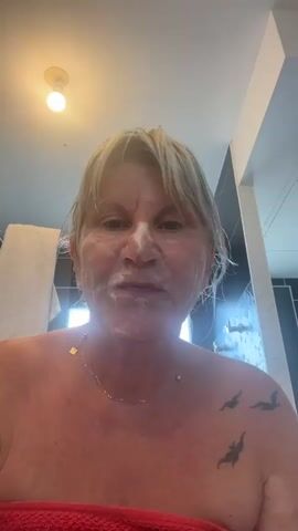The video shows a medium shot of a person with blonde hair, applying a white foamy substance to their face with their hands. The person's eyes are open and they are looking directly at the camera. Their mouth is slightly open, and they appear to be in the process of washing their face. The person is wearing a white shirt, and a red towel is visible around their neck. The background is out of focus but appears to be a bathroom with a light fixture on the ceiling. The person has tattoos on their left shoulder and arm. The video is approximately 4 seconds long.