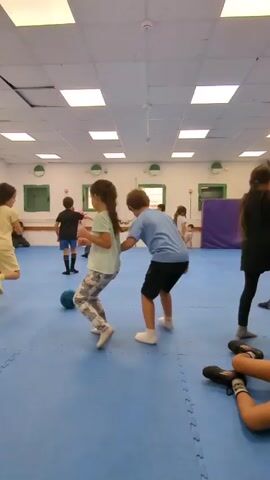 The video shows several children playing in a large, brightly lit room with a blue floor that appears to be made of interlocking foam mats. The room has a white tiled ceiling with evenly spaced fluorescent lights, and white walls. There are some windows along one wall and a purple object which looks like a padded gymnastics aid. There is a dark blue ball on the floor to the left of the frame. The children are of varying heights and are dressed in casual clothing. They are actively moving around the room and some of them are holding hands. Someone is speaking, likely giving instructions. One person is seen laying on the ground, on the right side of the frame.