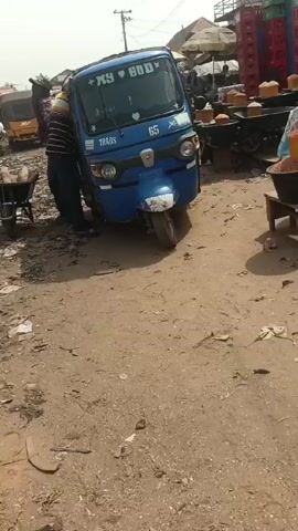 The video shows a blue tricycle parked in a crowded, dirty outdoor market. The tricycle has the words "MY GOD" on its windshield and the number "65" displayed on its front. A man in a striped shirt stands near the tricycle, possibly interacting with it. Beside the tricycle is a wheelbarrow filled with yams. The ground is covered in dirt and scattered debris. Other people can be seen walking around in the background, along with more tricycles and market stalls.