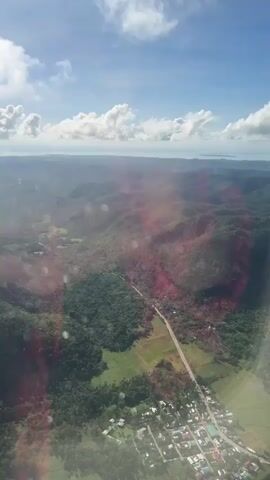 The view from an airplane window shows a landscape with rolling hills and dense forests under a bright blue sky with scattered clouds. A straight road cuts through the greenery, leading to a small town with closely packed buildings. The sun reflects off the window, creating a reddish glare and some light flares. The distant horizon appears to show the ocean.
