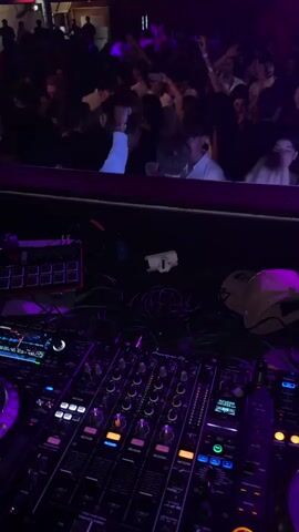 From a DJ's perspective, the video shows a crowded nightclub with people dancing under purple lighting and red balloons. The DJ is actively mixing music on a Pioneer DJ console, which is surrounded by tangled cables and other equipment. The club appears to be busy and lively.