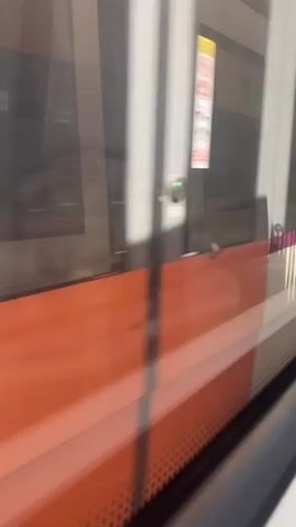 The video is a close-up, slightly shaky, interior shot of a train from the perspective of a passenger looking out the window. The window is large and reflects the interior of the train. The exterior of the train is visible as a white stripe with a thin purple stripe above it and an orange stripe below it. The train appears to be moving. The reflections in the window show the interior of the train, including seats and other passengers, but they are blurred and indistinct due to the movement and reflections.