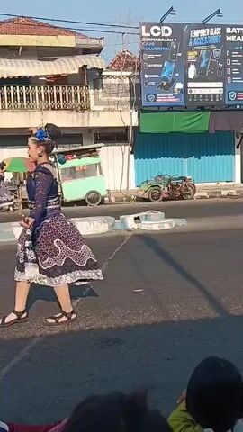 A young woman in a traditional blue and white patterned dress and elaborate blue hair accessory dances on a paved road. She is performing a graceful, slow dance with hand and arm movements. Her hair is pulled back in a bun. She is wearing sandals. The road is lined with buildings and shops on the right side of the frame. In the background, several other dancers in similar attire can be seen walking and dancing further down the road. A few motorcycles are parked along the sidewalk. People are seated on the side of the road, watching the performance. The sky is clear and blue, indicating a sunny day. There is a large billboard on the side of a building displaying advertisements for electronics. A green food cart is visible on the left side of the road.