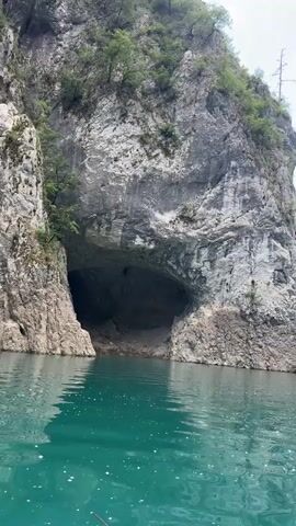 This media file captures a serene boat tour through a majestic canyon. The scene is dominated by towering, rugged cliffs on either side, covered in lush green vegetation, with a prominent cave opening visible on the left cliff face. The water is a striking turquoise, reflecting the sky and surrounding cliffs, with gentle ripples disturbing its surface.

The activity is clearly a boat journey through the canyon. In the distance, several small boats can be seen, some with people aboard, suggesting it's a popular tourist activity. The weather appears to be fair and sunny, with scattered white clouds in a bright blue sky. The time of day seems to be late morning or early afternoon, given the bright sunlight.

Notable details include the sheer scale of the cliffs, the deep turquoise hue of the water, and the ancient, almost prehistoric feel of the landscape. The presence of the cave hints at the possibility of further exploration or local folklore. The overall emotion evoked is one of awe, tranquility, and a sense of adventure.

While there is no visible text in the media, the location context provided is Bezuje, Montenegro, which helps to identify this as a scenic natural wonder within that region.
