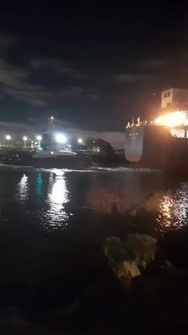 The video captures a nighttime scene at a dock in Dock Sud, Argentina. The primary object is a large cargo ship, its lights illuminating the dark water. The ship appears to be docked or maneuvering, with activity suggested by the illuminated decks and the wake in the water. The setting is a waterfront area, with a dark sky suggesting it is late at night. The water is reflective, mirroring the lights from the ship and other distant lights, creating a shimmering effect. On the left side of the frame, green and white lights are visible, possibly from navigational markers or dockside infrastructure. In the foreground, there is dark ground, possibly a quay or embankment, with some vegetation visible. The overall mood is industrial and somewhat serene, typical of a port at night. There is no clear indication of people, but their presence is implied by the ship's operation. No legible text is visible on the ship or in the surrounding environment.