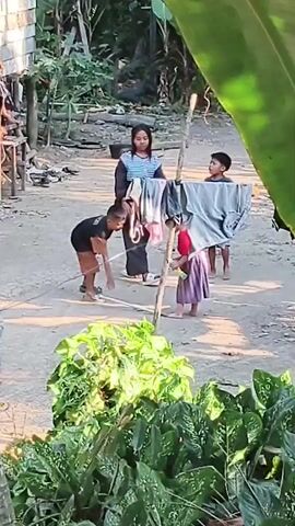 A group of children and one adult are outdoors in a dusty area with some structures and vegetation. The adult, a woman wearing black pants and a striped shirt, is walking towards a clothesline with laundry hanging on it. A boy in a black t-shirt and brown shorts is standing to her left, and another boy, wearing a light-colored shirt and shorts, is to her right, looking at the clothesline. A younger child in a purple dress is standing behind the clothesline. In the foreground, a young boy in a blue and white shirt is visible. The adult appears to interact with the children, and one of the boys is holding a rope.