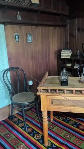 The video pans across a dimly lit room with wood-paneled walls. In the foreground, a wooden desk with carved legs is covered by a colorful striped rug. On the desk, there are several objects, including a dark glass bottle with a stopper, a small wooden box, a framed picture, and an oil lamp. To the left of the desk, a black metal chair with a woven seat is visible. In the background, a wooden shelf unit holds books and a green ceramic pitcher. Further to the right, a bed with red bedding and a fur throw is partially visible. Several framed pictures hang on the walls. The overall impression is of a rustic, old-fashioned living space.