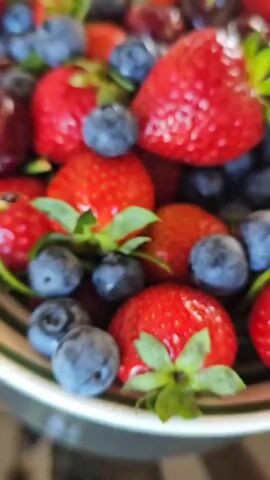 The video opens with an overhead shot of a white bowl filled with fresh red strawberries, dark blue blueberries, and dark red cherries on a dark, reflective surface. The camera slowly pans upwards from the bowl of fruit to a clear, patterned glass vase holding a bouquet of vibrant fuchsia-pink peonies with green leaves. The camera then zooms in slightly, focusing on the intricate petals of the peonies.