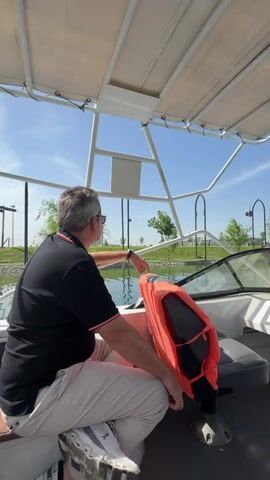 The video captures a serene daytime boat ride on a clear, sunny day in Kuchtumgali, Uzbekistan. The scene is set aboard a white recreational boat featuring a light-colored canvas canopy overhead.

Two individuals are visible in the boat. Initially, a man with short grey hair and sunglasses, dressed in a black polo shirt and light-colored pants, is seated on the left side, facing forward with his back partially towards the camera. He extends his right arm, appearing to point at something ahead or along the water's edge. An orange life jacket is draped over the boat's steering console, which also features a steering wheel and various gauges. The man's white sneakers, bearing the text "ON Cloud," are visible in the foreground.

As the camera slightly pans, another person, seen from behind and wearing a white T-shirt, becomes visible on the right side of the boat. The boat is navigating a body of water, likely a canal or a lake, flanked by well-maintained green banks dotted with trees and distinctive decorative archway-like structures. In the background, a modern, tall building, resembling a skyscraper, rises above the landscape, indicating the proximity of an urban area.

The overall impression is one of a calm and enjoyable outing on the water, with passengers taking in the scenic views under a bright blue sky.