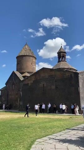 The video shows a group of people gathered in a courtyard in front of a stone building with a domed roof. The building appears to be an old church or monastery. The sky is blue with some white clouds. The ground is covered in grass and a stone pathway. A group of people, some dressed in formal attire, are standing and interacting. One person is walking across the grass. The lighting suggests it is daytime.