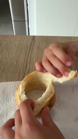 The video shows a child pulling apart a round, tan-colored bread or roll on a white paper towel on a wood-patterned surface. The child's hands are visible as they tear the bread. The background shows a stainless steel refrigerator and white cabinets. The child is wearing a white t-shirt with a cartoon graphic.