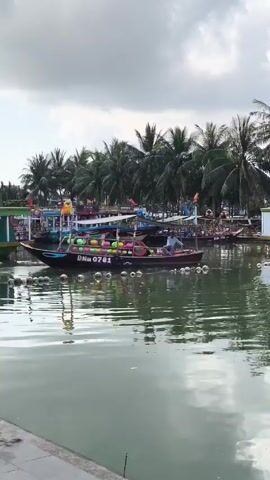 The scene shows a small river or canal with several boats docked along the bank. The water is calm, reflecting the sky and the boats. The sky is overcast with a layer of gray clouds. Palm trees line the bank, adding a tropical feel to the environment. One boat is in the foreground, loaded with colorful floats or buoys. A man is sitting in the boat, possibly the driver or operator. Other boats, some decorated with flags and colorful accents, are visible further back along the bank. Overall, the scene has a tranquil and somewhat overcast atmosphere.