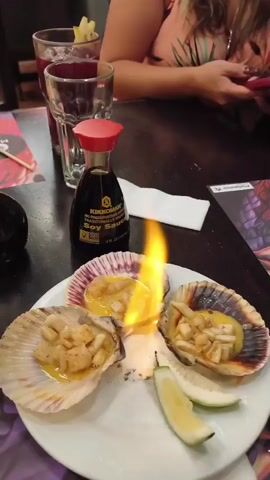The video shows a close-up of a white plate with three scallops in their shells, each filled with chopped seafood and a yellow sauce. Flames are rising from the center scallop. Two lime wedges are placed on the right side of the plate. A bottle of Kikkoman soy sauce stands behind the plate. In the background, there are two glasses with red beverages, a white napkin, and a person in a tropical-print shirt holding a phone. The table has a dark surface. The video appears to be taken in a restaurant setting.