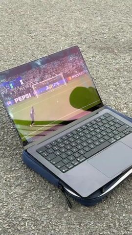 A soccer game is shown on a laptop screen. The laptop is set on the ground. A group of people are watching the game. A man with long curly hair is wearing a black and green soccer jersey. He expresses emotion regarding the game. He is sitting on the ground near a chain link fence.