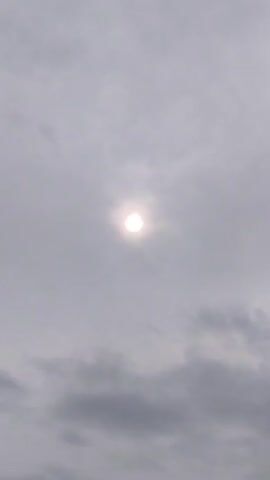 A video shows the sun partially obscured by clouds. The sun appears as a bright, white circle surrounded by a soft halo. The clouds are gray and diffuse, creating a hazy atmosphere. The camera appears to be stationary, and the sun remains in the center of the frame throughout the short clip.