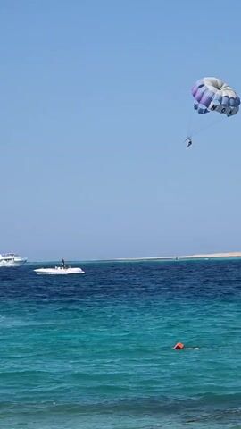 The video shows a parasailer suspended in the air by a parachute over a body of water. The parachute is multicolored with purple, white, and blue sections. Two figures can be seen attached to the parachute, presumably the parasailer(s). Below the parasailer, a jet ski is visible on the water, and further in the distance, a white boat is moving across the water. The sea is a deep blue, transitioning to turquoise closer to the shore, with visible waves. A small orange buoy is floating in the water. In the background, a strip of land with some vegetation can be seen. The sky is clear and blue.