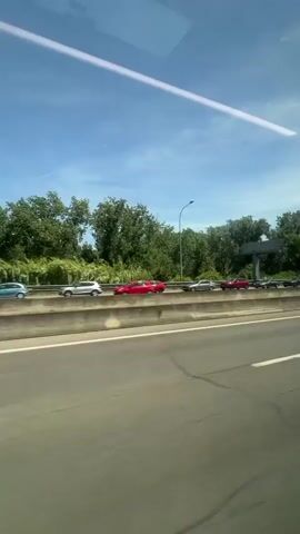 The video shows a view from a vehicle traveling alongside a highway. Cars are visible on the highway, moving in the same direction. The highway has multiple lanes and is bordered by a concrete barrier. Beyond the barrier, there are trees and a clear blue sky with some clouds. The sun is shining, casting shadows. There are no people visible. The overall scene depicts a typical highway environment on a sunny day.