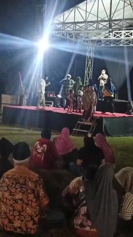 The video shows an outdoor nighttime performance on a raised stage. A bright spotlight illuminates the stage, creating lens flare. On the stage, several people are present, including performers dressed in various clothing. Some individuals appear to be speaking into microphones. In front of the stage, an audience is seated or kneeling on the grass, also facing the performance. The audience members are wearing diverse attire, including headscarves. The environment appears to be a grassy outdoor area with trees visible in the background. The video quality is somewhat grainy, and there are moments of motion blur, particularly in the latter half.