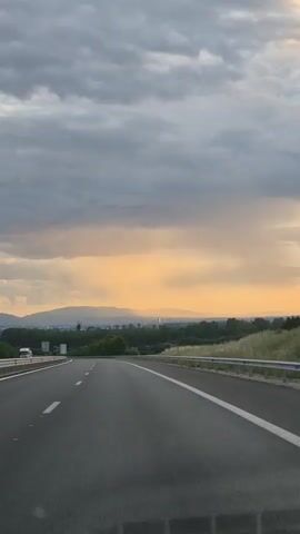 The video shows a view from a moving car on a highway. The sun is setting, casting a warm orange glow across the sky. Dark, heavy clouds dominate the upper portion of the frame. In the distance, there are rolling hills and a few distant buildings. The highway is empty except for a white truck far ahead on the left lane. The camera appears to be mounted on the dashboard or windshield. The road has clear white lane markings.