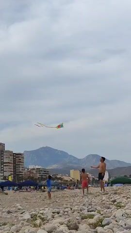A man and two children are on a pebble beach flying a kite. The man is wearing black shorts. One child is wearing a blue shirt and patterned shorts, and the other child is wearing red shorts. The kite is colorful and is flying in the sky. In the background, there are buildings, tents, and mountains. The sky is cloudy.