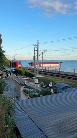 The media depicts a coastal scene in Khostinskiy, Russia, likely during late afternoon or early evening due to the warm, golden light and long shadows. The sky is a vibrant blue with scattered, wispy clouds, indicating pleasant weather.

The main subjects are a bright red and silver passenger train moving along tracks adjacent to the Black Sea. The train has "Ласточка" (Lastochka) written in red on its side, which translates to "Swallow," a common name for this type of electric train in Russia.

In the foreground, there's a raised walkway with a metal railing, overlooking a green, landscaped area. This area appears to be a leisure or public space with several people visible. Some are standing and observing, while others are seated or lying down on lounge-like structures, suggesting relaxation or recreation. There are also some colorful, abstract-shaped structures that might be artistic installations or play equipment. A large, gray, corrugated metal roof dominates the very bottom of the frame, indicating some form of building or structure.

The setting is clearly a seaside location, with the calm blue water of the Black Sea stretching to the horizon. A light-colored building with a red roof is situated between the train tracks and the sea, possibly a station or a cafe. The presence of palm-like plants in the foreground suggests a subtropical climate.

The overall atmosphere seems tranquil and summery, with people enjoying the outdoors near the coast while a train passes by. The activity involves leisurely lounging and observing the surroundings, with the train's movement adding a dynamic element to the scene.