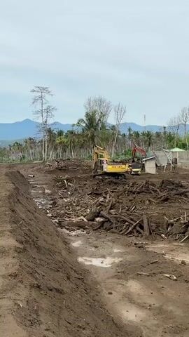The media depicts a scene of post-disaster recovery and rebuilding efforts in Meunasah Lhok, Indonesia. The foreground is dominated by a muddy, debris-strewn landscape, suggesting significant damage from a natural event, likely a flood or landslide, given the abundance of uprooted trees and logs. A trench or channel has been dug on the left side of the frame, filled with muddy water.

In the mid-ground, heavy machinery, including a yellow excavator and a red backhoe, are actively engaged in clearing operations. A pile of logs and debris is being worked on by the excavator. To the right, construction is underway on several structures. A yellow building with a red roof is partially visible, and a smaller white building with a green roof is also present. Several yellow dump trucks are parked in the vicinity, implying ongoing transportation of materials or debris. Two figures, possibly workers, are visible near the construction site.

In the background, a dense line of palm trees stretches across the horizon, interspersed with some bare trees. Beyond the trees, mountains are visible under a cloudy, overcast sky, suggesting it is daytime, likely morning or afternoon, with no direct sunlight. The weather appears to be humid, typical for the region. A tall telecommunications tower stands in the distance.

The overall impression is one of arduous work and the slow process of reconstruction in the wake of a disaster. The scene conveys a sense of resilience and ongoing efforts to restore the community. There is no visible text in the media.