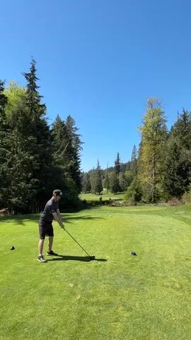 A man is teeing off on a golf course on a sunny day. He is wearing a gray shirt, black shorts, a black baseball cap, and athletic shoes. He is holding a golf club and swinging it to hit a golf ball. The golf ball is on a tee in the middle of the frame. The course is surrounded by trees, and the sky is clear and blue. There are two additional tees on the ground near the golfer, one blue and one black.