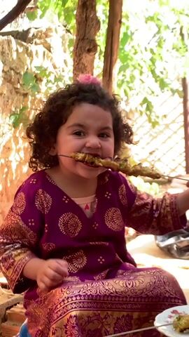 Here is a brief description of the video:
A young girl with curly hair and a pink flower in her hair sits outside, cross-legged, wearing a purple and gold patterned dress. She holds a metal skewer with grilled meat, taking a bite, and then smiles at the camera. The background features greenery, brickwork, and dappled sunlight.