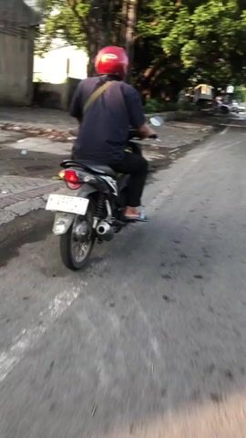 A person wearing a red helmet and a dark blue shirt rides a black motorcycle on a paved road. The person's back is to the camera, and they are wearing black pants and flip-flops. A strap from a bag is visible across their shoulder. The motorcycle has a white license plate with black lettering. The road appears to be in an urban or suburban area, with some vegetation and a fence visible in the background. The lighting suggests it is daytime, possibly late afternoon or early morning due to the direction of the shadows. The camera is positioned slightly behind and to the side of the motorcycle, capturing its movement.