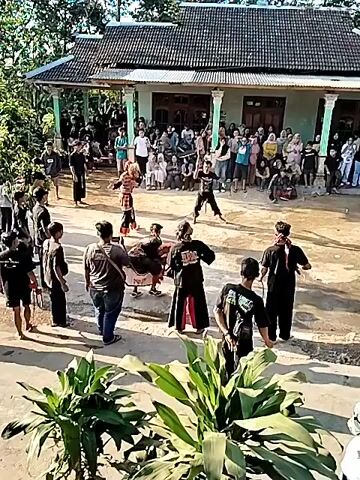 A group of people are gathered in a courtyard in front of a building, watching a performance. Several individuals are actively participating in the performance, some wearing traditional clothing and masks, and are engaged in what appears to be a martial arts or dance demonstration. One performer is on the ground, while others are standing or moving around. There are spectators sitting and standing around the perimeter of the performance area. The environment is outdoors, with a house in the background and vegetation in the foreground. The overall atmosphere suggests a cultural event or festival.