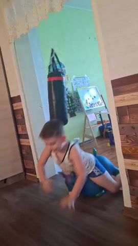 A young child is on the floor in a room with a punching bag hanging above. The child is lying on their back and appears to be struggling or playing with an inflatable blue toy. The punching bag is black with red accents and has an image on it. The room has a light green wall and wood-paneled walls in the background. There is also a wardrobe on the left side of the frame and an easel with a drawing on it visible in the background. The floor is wooden. The child's body language suggests they are engaged in playful activity.