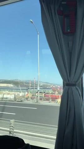 The video captures a scenic view from inside a moving vehicle, likely a bus, looking out a large window. A gray curtain is pulled back and tied, framing the right side of the window, and a red emergency tool is visible attached to the window frame. The sky is clear and blue, indicating daytime.

Initially, the view encompasses a large industrial port on the right, filled with numerous cranes, ships, and industrial structures. A multi-lane highway, possibly an overpass, runs in the foreground. As the vehicle moves, the scenery transitions. The port recedes into the background, and a vast expanse of turquoise sea comes into view, stretching towards distant hills or mountains on the horizon. A tall, white street lamp passes by in the mid-ground. The vehicle appears to be traveling on a bridge or an elevated road alongside the water. The audio in the background mentions "one of the new bridges in the country" and its length being "2000," which likely refers to meters, further confirming the location is on a significant bridge.