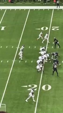 The video shows an American football game in progress, filmed from an elevated angle, likely from the stands. Two teams are lined up in a typical offensive-defensive formation at the 20-yard line of a football field. The players are wearing full football gear; one team is in white uniforms, and the other is in dark uniforms with green accents. The offensive team snaps the ball, and the quarterback drops back to pass. He throws the ball downfield. The background shows the green field with yard markers and the outlines of other field markings. Spectators can be seen in the blurry background around the field.
