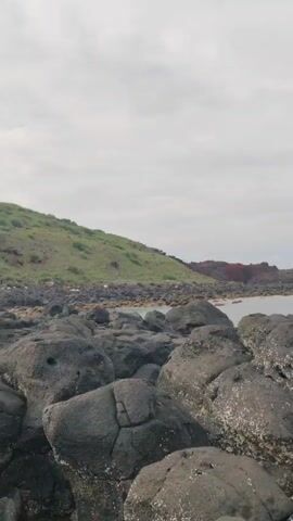 The video shows a rocky beach with a small grassy hill and a body of water, likely the ocean, on a cloudy day. The foreground is filled with large, dark gray rocks covered with white lichen. In the midground, there is a grassy hill to the left and a reddish-brown rocky area extending into the water to the right. The water appears calm and gray. A person with long, dark hair wearing a light gray long-sleeved shirt walks into the frame from the left, briefly appearing in the midground before disappearing to the right.