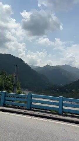 The video captures a view from a moving vehicle on a road, likely a bridge, looking out at a mountainous landscape. In the foreground, a bright blue railing with white horizontal bars runs along the side of the road. Two tall, grey poles with numbers "4" and "5" are visible on the railing, along with a yellow "NO PARKING" sign. Beyond the railing, a wide, muddy river flows through a valley. Steep, lush green mountains covered in trees rise on either side of the river. The sky above is partly cloudy with bright sunlight filtering through. In the distance, nestled on the hillside, are several buildings, some appearing to be hotels or resorts, with white walls and green roofs. The perspective suggests the vehicle is moving forward, offering a panoramic view of the scenery.