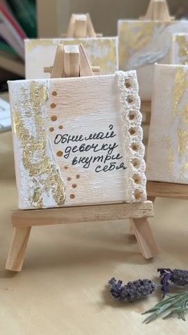 The video shows a group of small, handmade artworks displayed on tiny wooden easels. Each artwork is a square canvas, decorated with a cream and white background, gold leaf accents, and handwritten text in black ink. Some of the canvases also feature delicate embellishments like crochet lace or small bows with dried lavender. The artworks are arranged on a light-colored surface, with sprigs of dried lavender scattered around. The camera pans slowly across the artworks, showing the different designs and messages on each one. The lighting is soft and natural, highlighting the textures and details of the artwork.