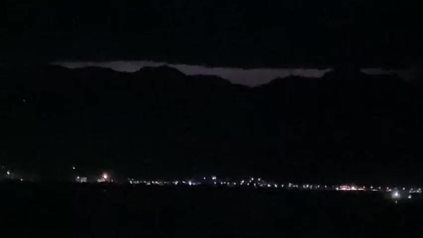 This is a video of a nighttime cityscape with mountains in the background. Lights from buildings and streetlights are visible in the city. The sky is dark with some clouds. There is a faint glow in the sky above the mountains, possibly from lightning or distant city lights. The video is dark and grainy, suggesting it was filmed in low light conditions. The footage appears to be static, with no visible camera movement. There are no people or objects clearly discernible in the foreground.