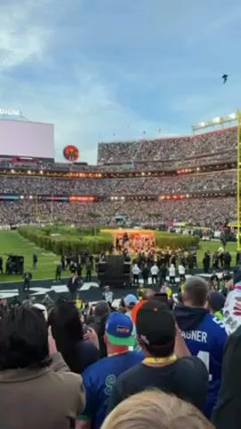 The video shows a large crowd filling a stadium, presumably for a sporting event or concert. The stadium is packed with people in the stands. On the field, there appears to be a performance or presentation taking place, with a stage and several people standing on it. In the foreground, viewers are seen with their backs to the camera, recording the scene. Some are clapping and raising their arms. Fireworks are launched above the stadium as well. The sky is partly cloudy.