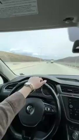 The video shows a view from the driver's seat of a car driving on a highway. The driver, wearing a watch and rings, has their left hand on the steering wheel.  The highway has multiple lanes and is flanked by hills and mountains in the distance.  There are streetlights along the side of the road. Other vehicles can be seen further ahead on the highway. The sky is overcast.