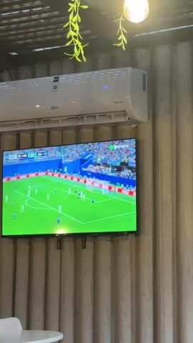 The video shows a television screen mounted on a wall displaying a soccer match. The television is positioned below a white air conditioning unit. The wall is made of corrugated metal. Above the television, there are some green plants hanging from a slatted ceiling. The soccer match shows players in white and blue uniforms on a green field with spectators in the background. The air conditioning unit displays the number 24. In the foreground, a white table and chair are partially visible.