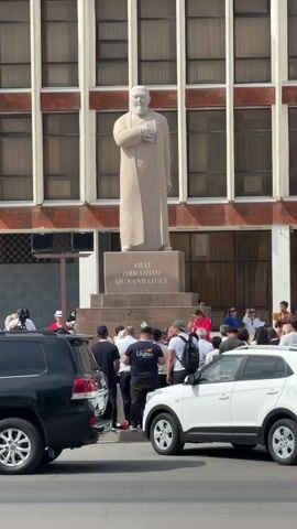 The video shows a statue of a man in a long robe and beard standing on a pedestal in front of a building with many windows. Several people are gathered around the base of the statue, some are taking pictures, and others are talking. There are cars in the foreground, a black SUV on the left and a white SUV on the right. The overall scene appears to be an outdoor public space with a monument.