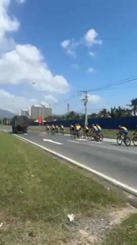 The video shows a group of cyclists riding in formation on a paved road. The cyclists are wearing matching jerseys with blue, yellow, and red colors. They are riding on road bikes. A military truck is seen further ahead in the same lane. In the background, there are tall buildings, a construction crane, and mountains in the distance. The sky is blue with some clouds. Grass lines the side of the road. A motorcycle can be seen driving alongside the cyclists. There's a large banner on the side of the road in front of the buildings.