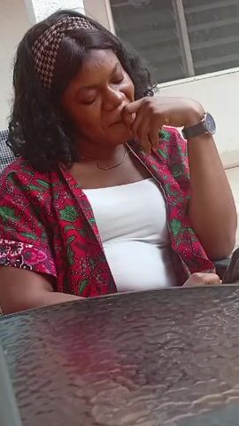 A woman sits at a glass table, wearing a white top and a red and green patterned shirt. She has a patterned headband on her head and a watch on her left wrist. She rests her chin on her left hand and looks to the right, then down. She appears to be thinking or contemplating something. The background includes a white wall and a window.