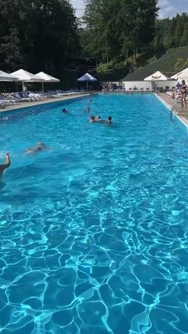 The video shows a long, rectangular swimming pool filled with bright blue water. Several people are swimming in the pool. In the foreground, a boy is swimming towards the camera. Further back, a group of children and adults are spread out in the pool. On the right side of the pool, there are several people standing and watching. The pool is surrounded by lounge chairs and umbrellas. In the background, there are trees and a hillside. The sky is clear and the sun is shining.